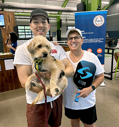 Two people smile holding a dog and pickleball paddle