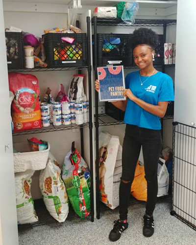 A young woman holds up a sign saying Pet Food Pantry in front of a closet used to store pet food.