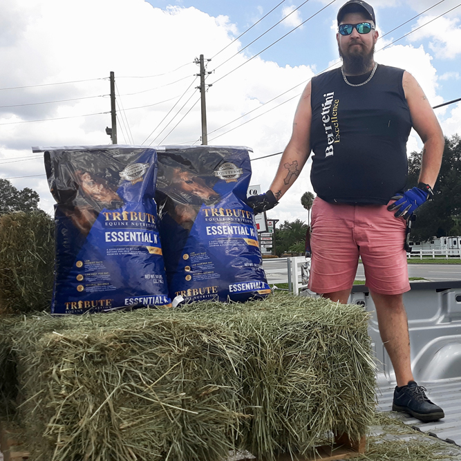 a man standing alongside several bales of hay and bags of horse feed