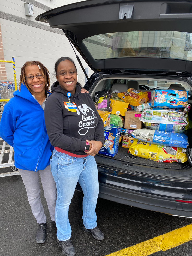 two women pose in front of a car trunk full of pet food and supplies