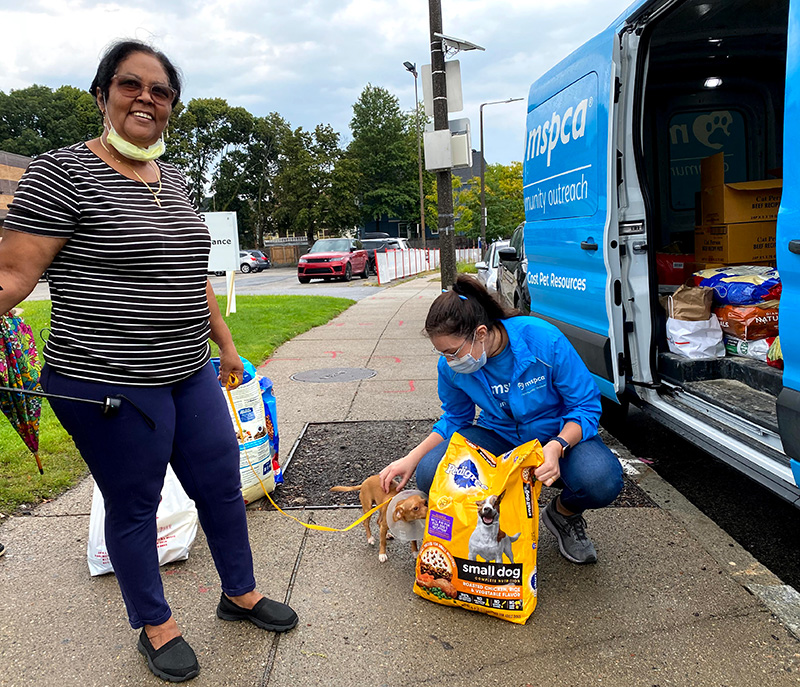 a shelter worker pets a small dog as she hands the dog's owner a bag of pet food