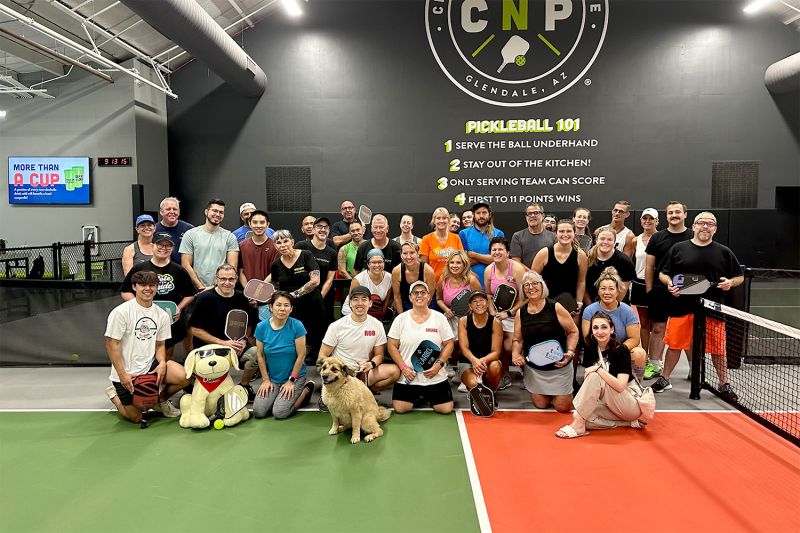 a large group of people pose with a dog on a pickleball court