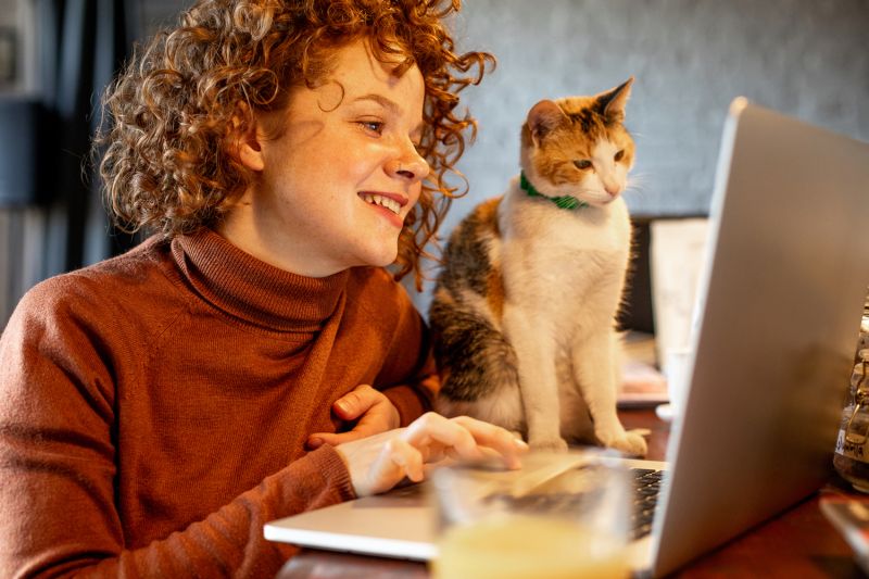 a woman smiles at her laptop screen while her cat looks on