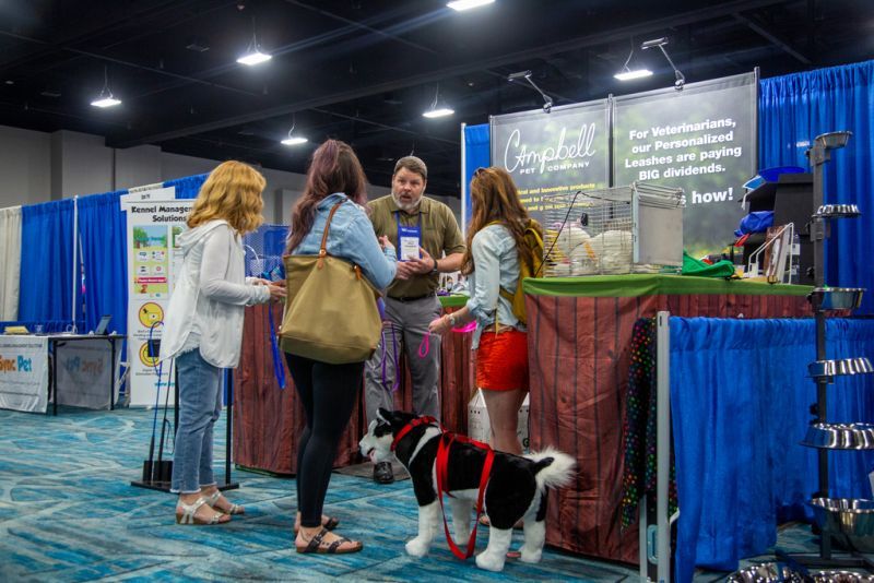 a man speaking with a group of people at a booth in the exhibit hall