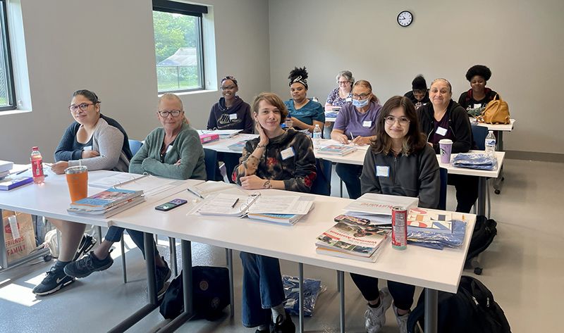 A group of people sit at tables in a classroom setting.