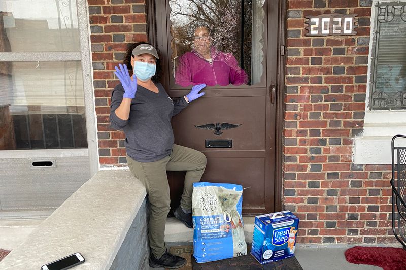 a woman with pet supplies waves from a porch while the owner waits within the house