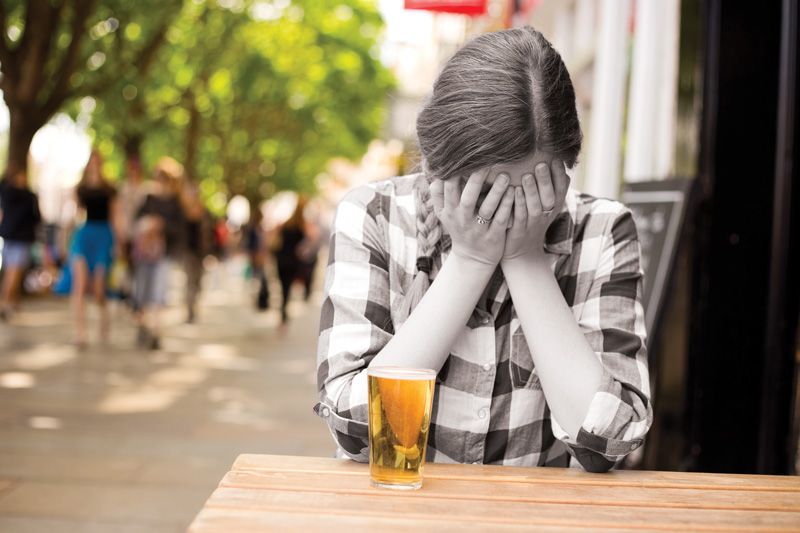 a woman sits with her face in her hands next to a glass of alcohol