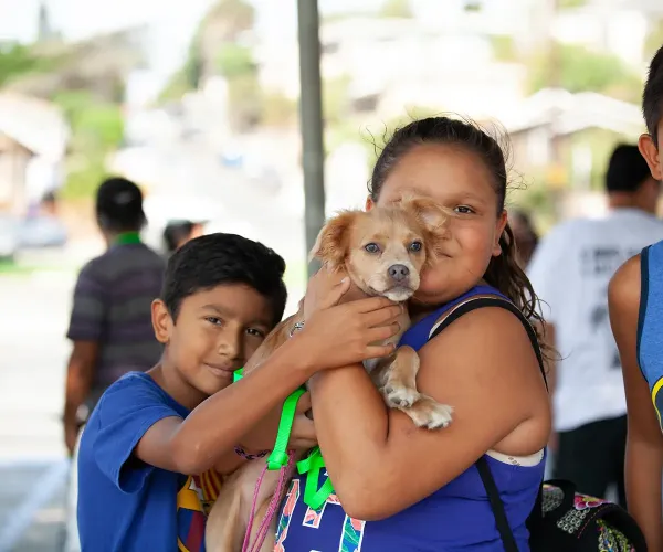 Three children smiling while holding a tan dog together at an outdoor community event, wearing blue shirts and colorful wristbands.