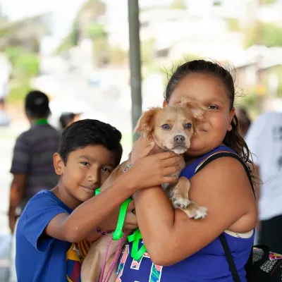 Three children smiling while holding a tan dog together at an outdoor community event, wearing blue shirts and colorful wristbands.