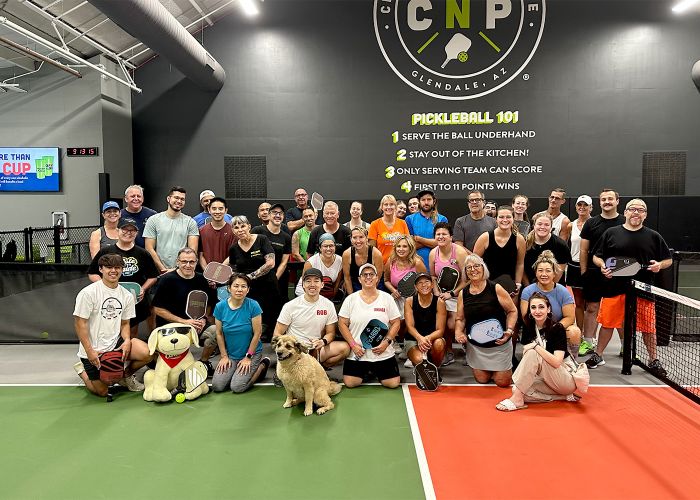 a large group of people pose with a dog on a pickleball court